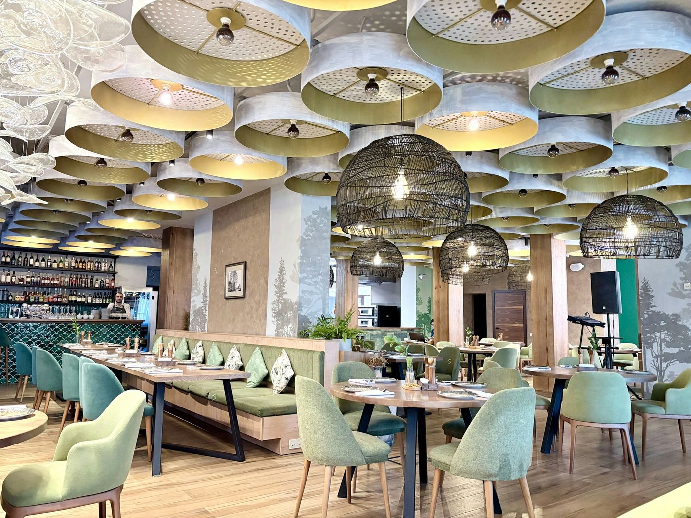 A bright restaurant dining room with sage-green chairs, set tables and woven pendant lights