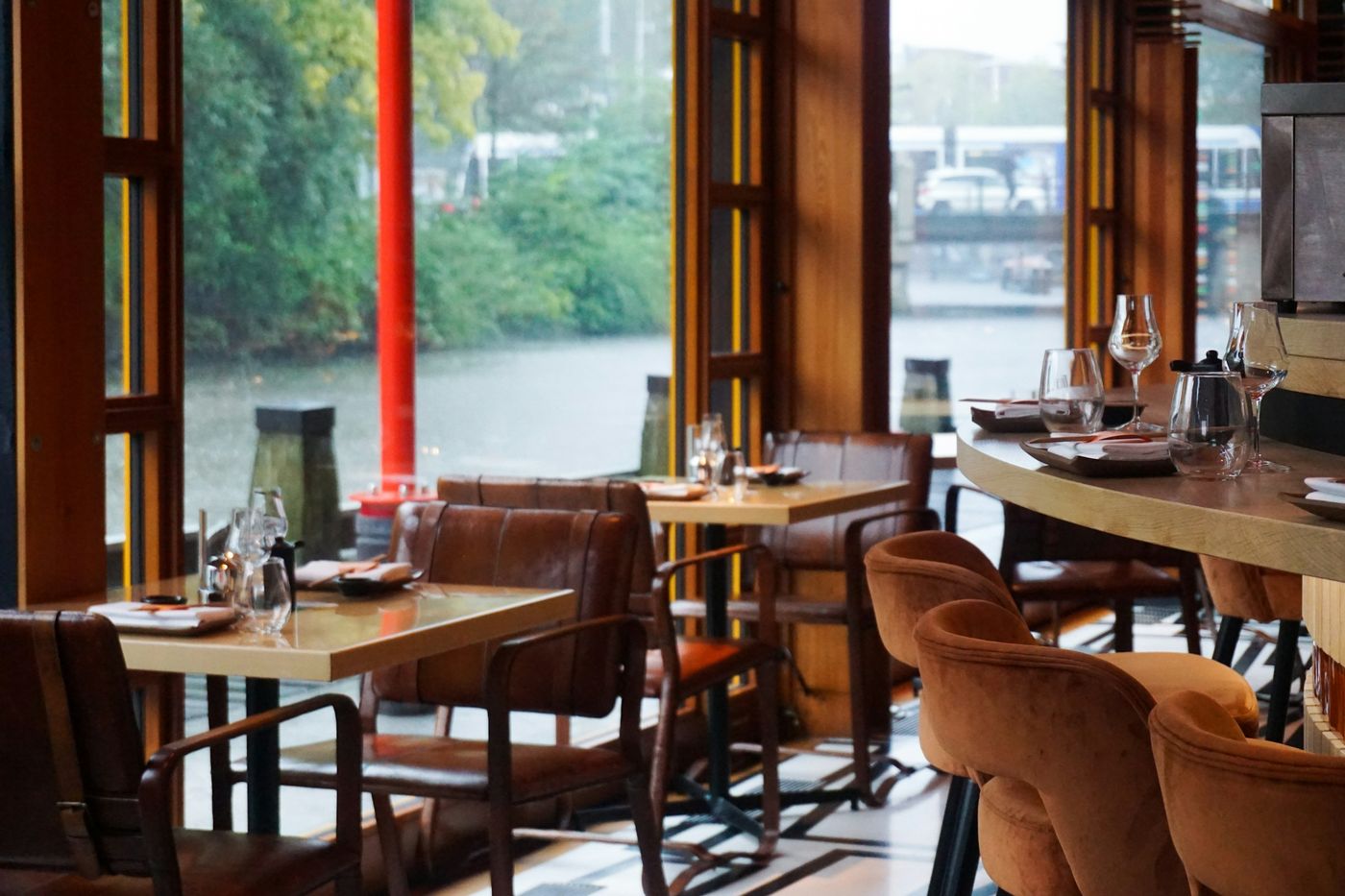 A restaurant with leather chairs and set tables beside tall windows onto the water