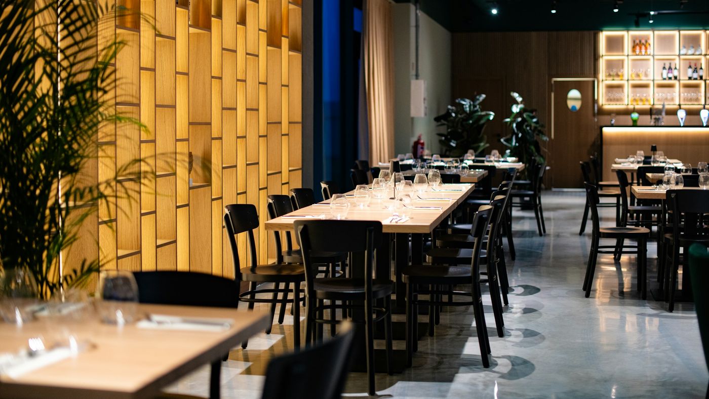 A restaurant dining room with long set tables, black chairs and a lit wooden wall