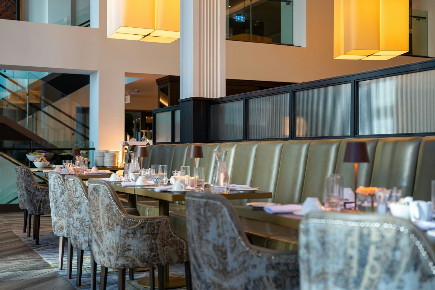A hotel restaurant with set tables, a long leather banquette and large pendant lights
