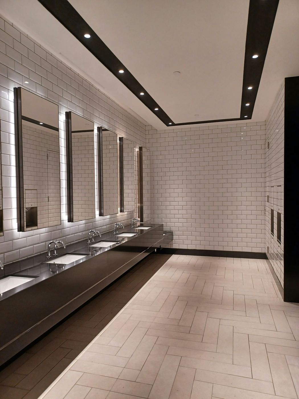 A new restroom with a long dark vanity, lit mirrors, white tile and a herringbone floor